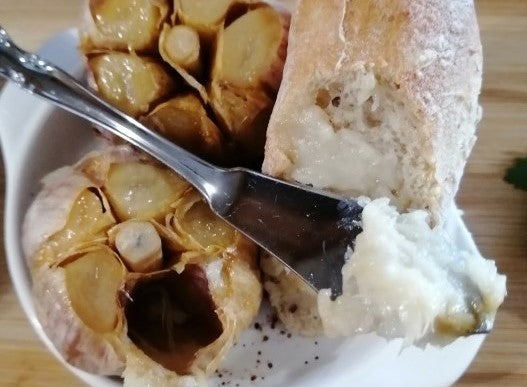 Roasted garlic being spread on a baguette with 2 whole roasted garlic bulbs on the plate beside the baguette